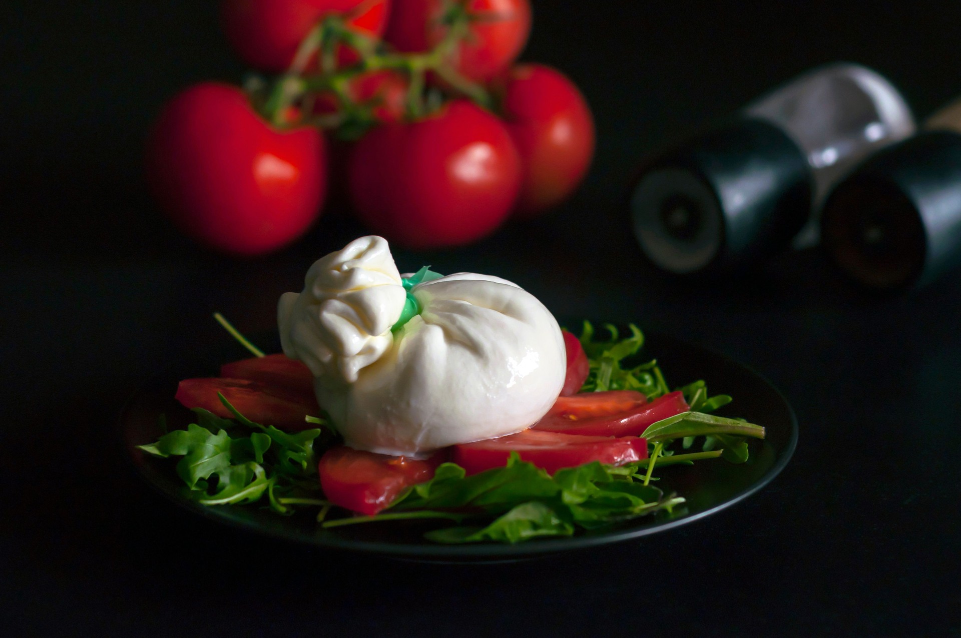 on a black plate is a salad of spinach and arugula greens, sliced tomatoes and Italian burrata cheese, in the background you can see a bunch of red tomatoes and spice mills, side view.